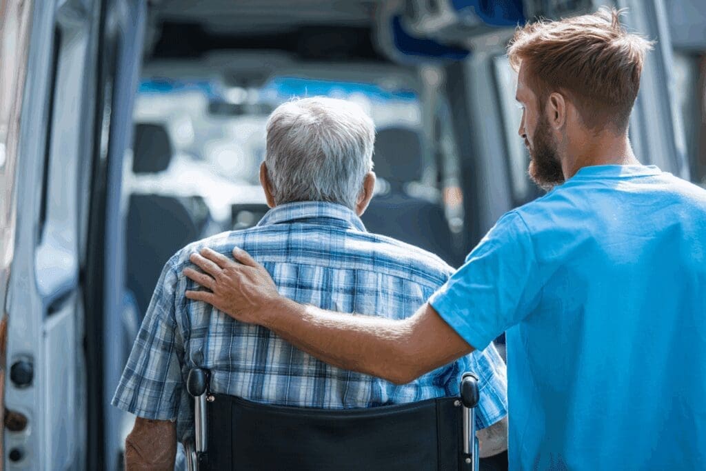 A caring transport driver in sky blue t-shirt gently placing a hand on the shoulder of an elderly patient in a wheelchair, guiding them into a medical van. Warm, empathetic scene. 