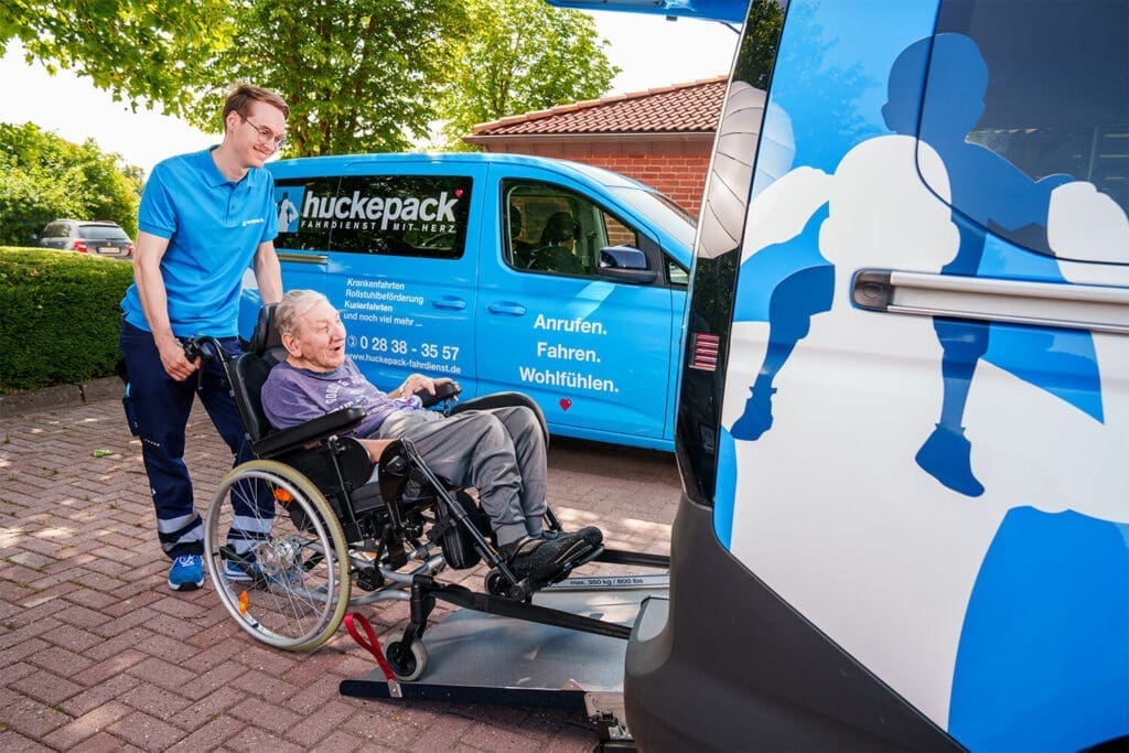 Ein älterer Mann im Rollstuhl wird fürsorglich von einer Huckepack-Mitarbeiterin ins Fahrzeug begleitet – Mobilität mit Herz und Respekt.